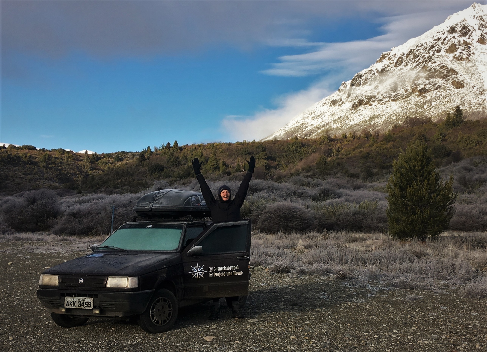 Tarcísio comemorando ao lado do Fiat Uno motorhome na Patagônia com montanha nevada ao fundo